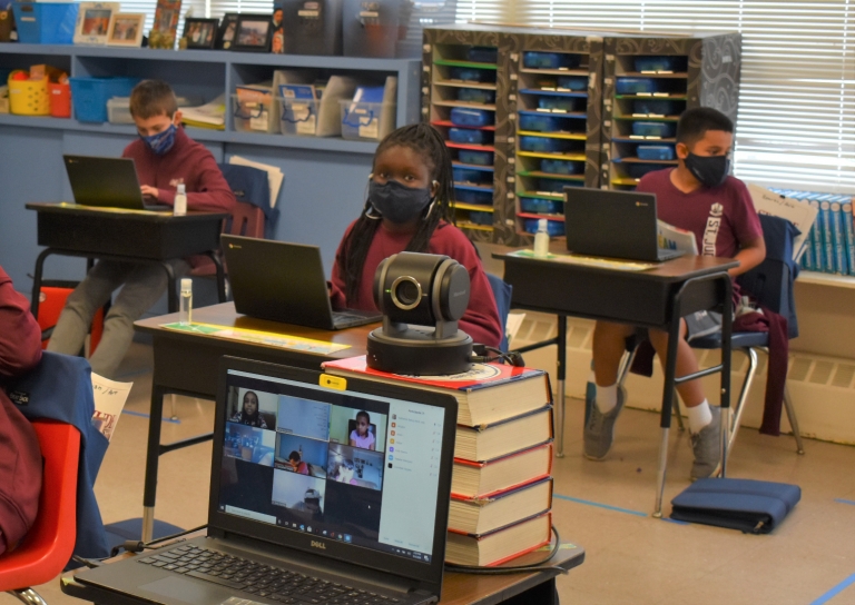 Fourth grade students use Chromebooks, foreground shows classmates joining virtually via Zoom