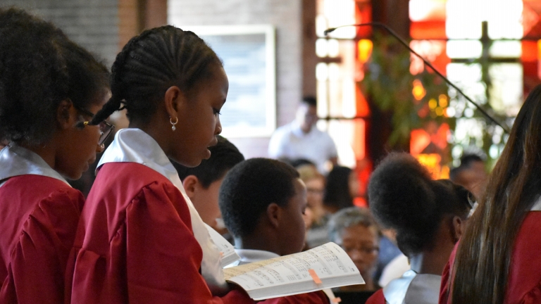 A sixth grade student sing from a hymnal with her friends with stained glass in the background
